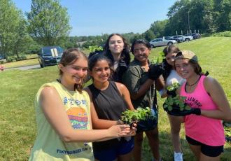 kids standing in grass field wile holding potted plants with cars in the background