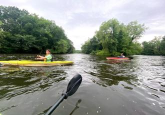 Two kayakers paddle on a calm river surrounded by lush greenery under a cloudy sky.