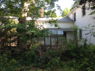 A partially obscured house surrounded by dense greenery and trees, featuring a glass-enclosed porch.
