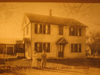 A vintage photograph of a two-story house with a porch, featuring two women standing in front. The image has a sepia tone.