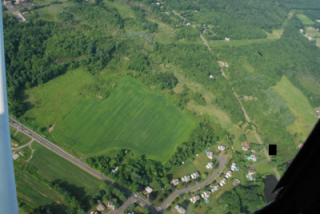 Aerial view of green fields and residential areas, with a road running through the landscape. Lush trees surround the area.