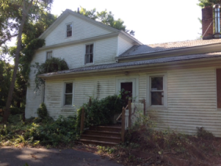 An old, two-story house with white siding, overgrown plants, and a wooden staircase leading to the front door.